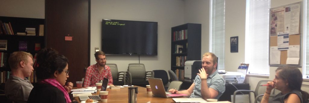 A few people chat while sitting at a conference table. Full bookshelves are in the background.