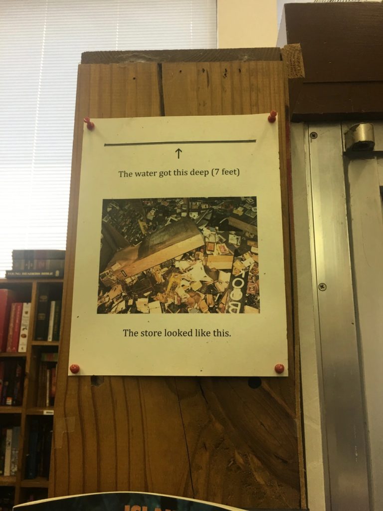 Photo of a piece of paper tacked on the side of a wooden bookshelf. The paper says “the water got this deep (7 feet). The store looked like this.” On the paper is an image of materials in disarray.