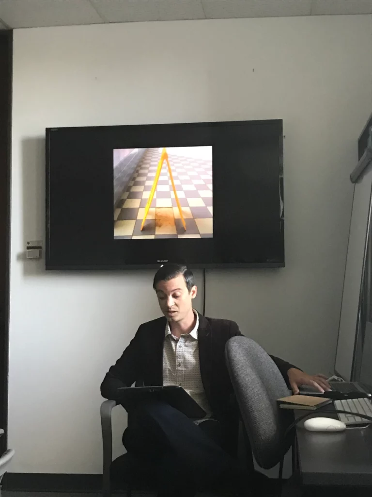 A person reads aloud from notes while sitting in front of a screen showing an image of a wet floor caution sign.