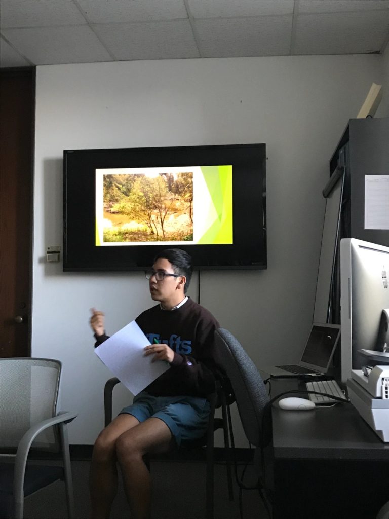 A person speaks while sitting in front of a screen showing an image of trees.