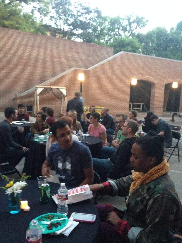 People sit at round tables with black tablecloths in a brick courtyard. There are flowers and plates of food on the tables.