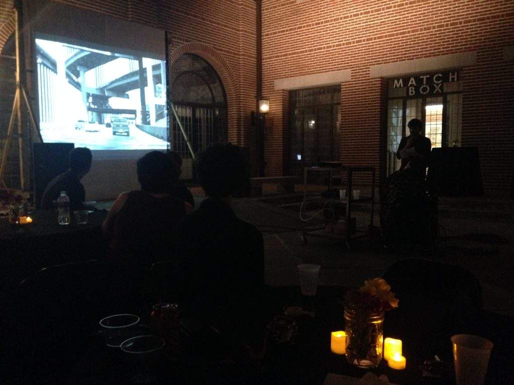 A projected slide is illuminated in a red-brick courtyard at night. Candles and flowers are arranged at tables in front of the projector.
