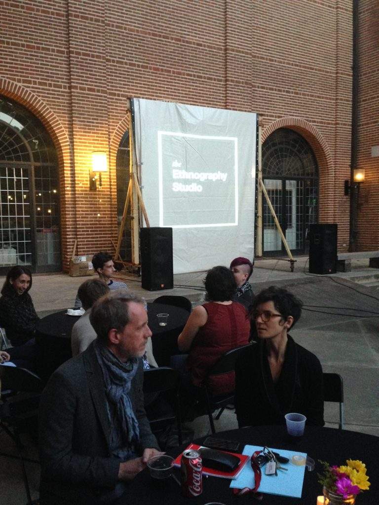 People chat while sitting around tables with black tablecloths in a brick courtyard. In the background, the Ethnography Studio is projected onto a draped screen.