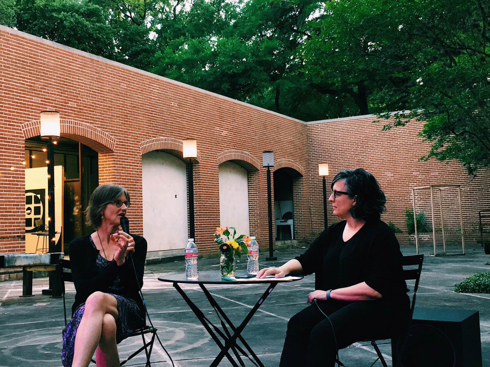 Two people sit in conversation, with a microphone, at a small metal table in a brick courtyard.