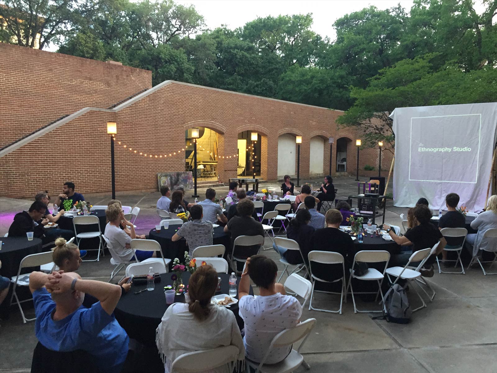 Several dozen people sit at round tables in a red-brick courtyard. At one end of the courtyard is a screen onto which the Ethnography Studio logo is projected.