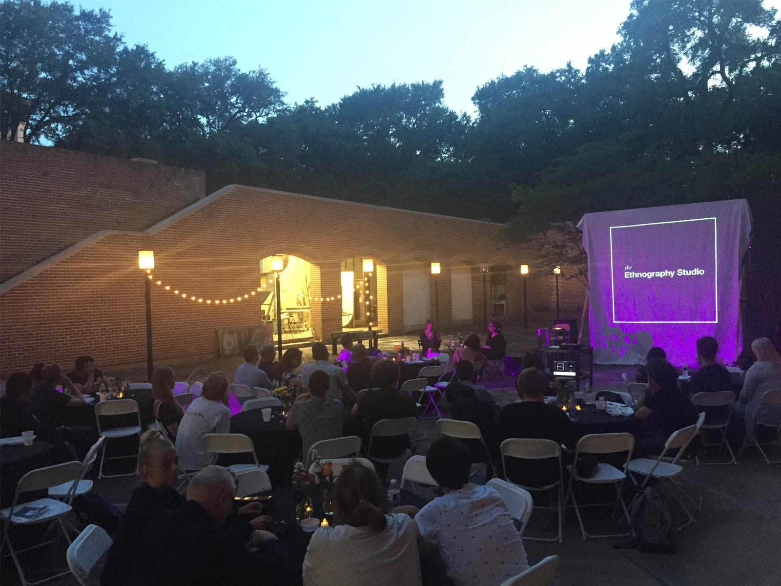 Several dozen people sit at round tables in a red-brick courtyard. At one end of the courtyard is a screen onto which the Ethnography Studio logo is projected. It is near dusk and the fairy lights strung up on one side are bright.