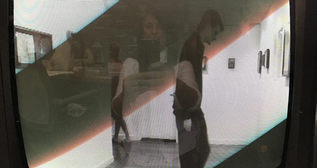 A photo of an old TV box displays an image of someone standing in an art gallery. Barely perceptible in the screen is the reflection of the person who took the photo.