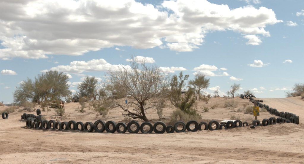 Photo of a row of rubber tires propped up as a fence in a desert landscape, with light tan sand, shrubby trees, and a light blue sky with fluffy clouds.