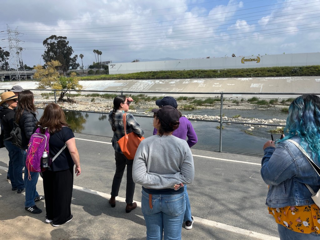 A handful of people stand in front of a railing along a concrete embankment along a shallow river, looking at the scene.