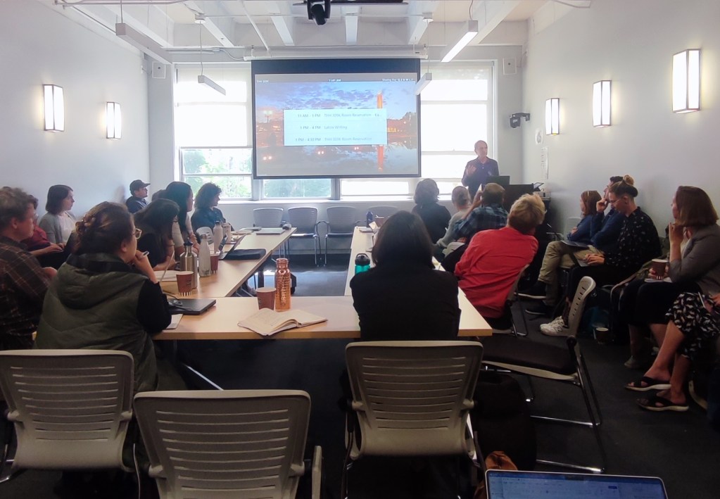 About a dozen people sit around a u-shaped conference table, listening to a speaker who stands in front of a projected slide.