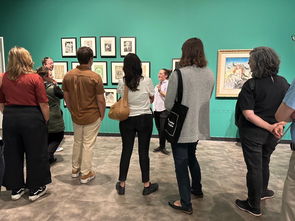 A handful of people stand with their backs to the camera, listening to someone speaking while standing in front of an art museum wall.