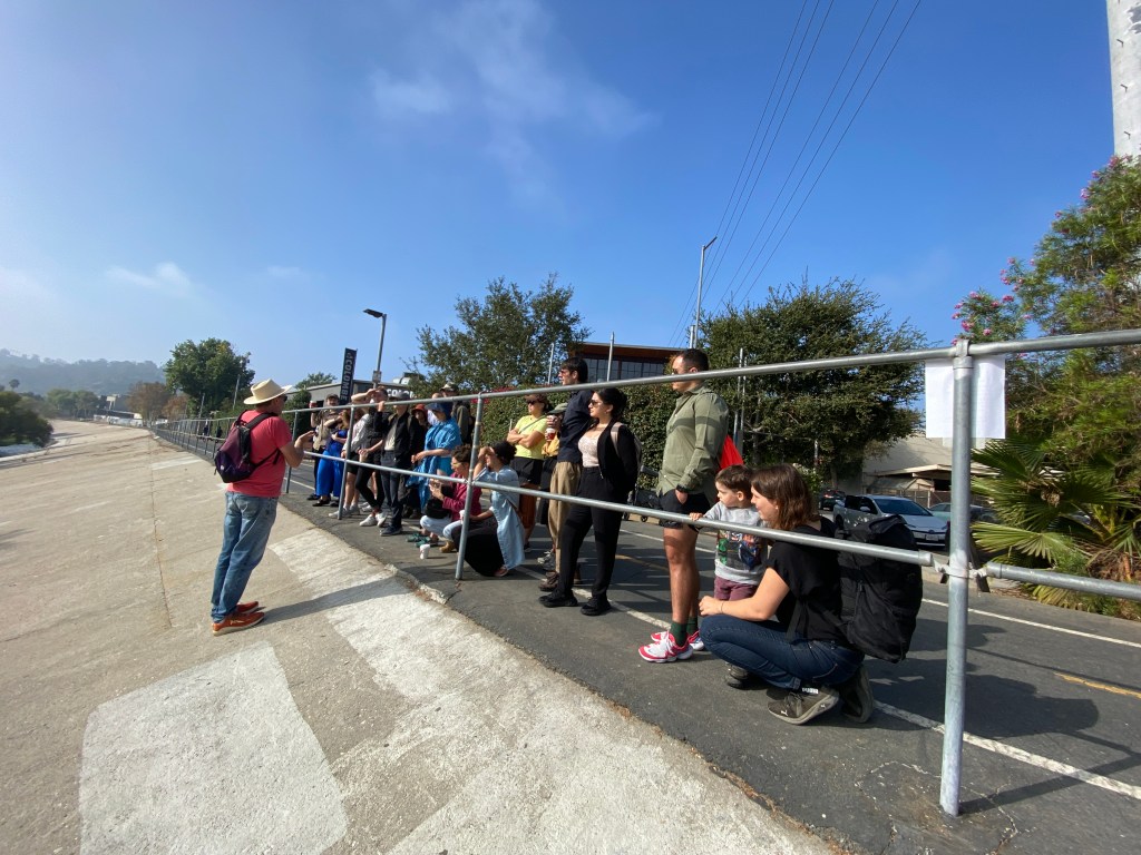 A group of about a dozen people stand behind railings listening to someone on the other side of the railing, on a gently sloping concrete embankment. On the other side of the group of people is a bicycle path and trees.