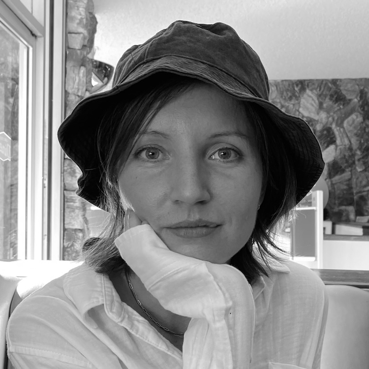 Black and white headshot of a person with a bucket hat, softly smiling, chin-length straight hair, and a light-colored linen button-up shirt.