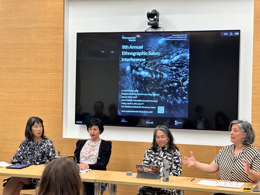 Photo of four people sitting at a table, presenting to an unseen audience. Behind them is a screen showing a slide with a poster for the event.
