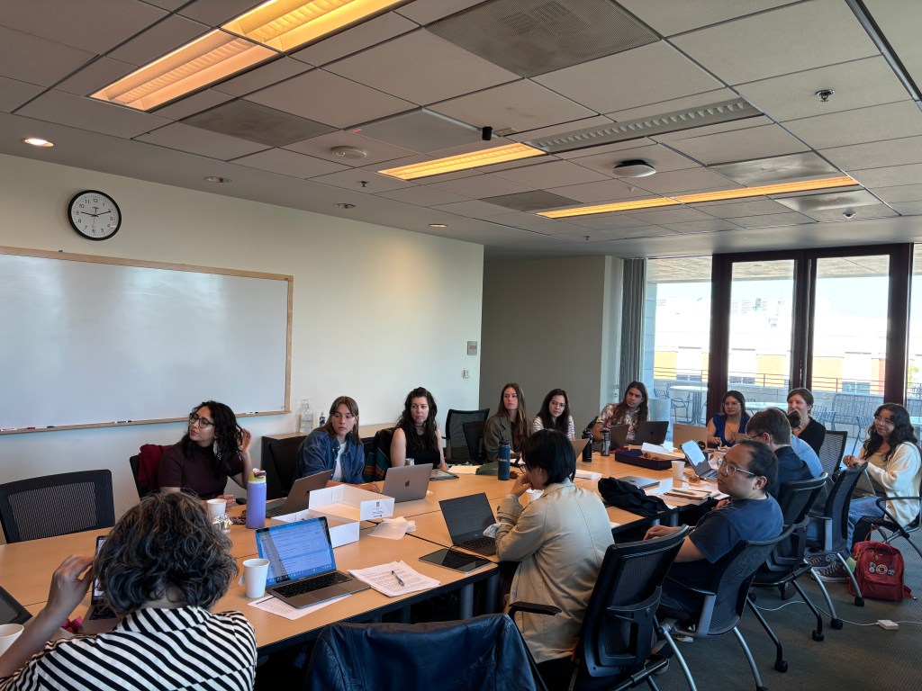Over a dozen people sit around a conference table in a spacious room. One end of the room is floor-to-ceiling windows and bright light pours in. Most people have laptops in front of them but look towards the front of the room toward an unseen speaker.