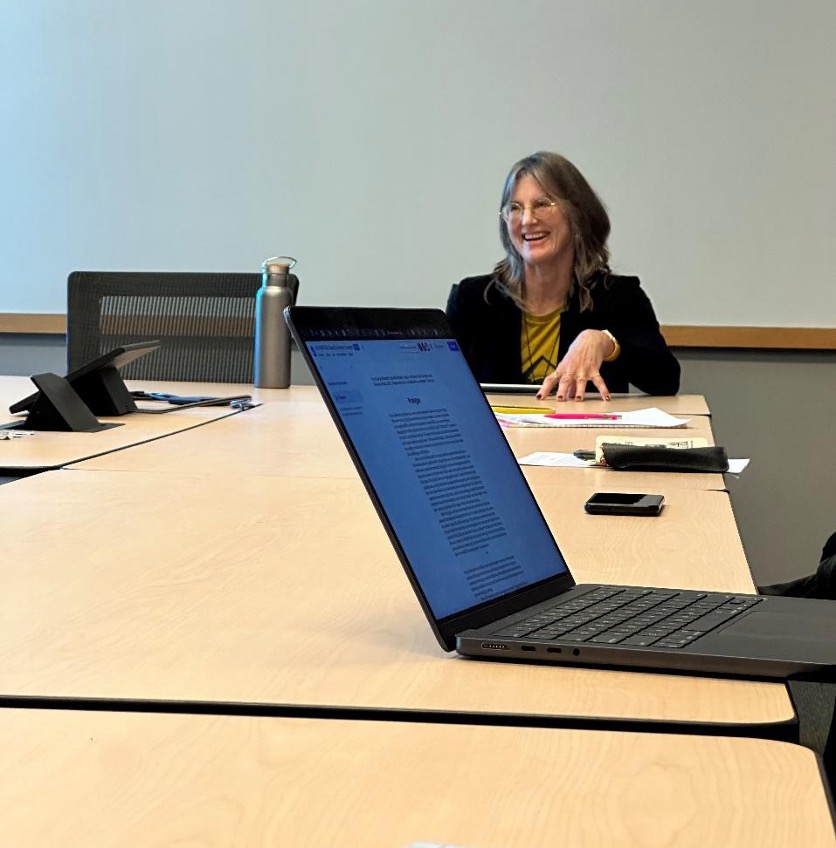 Someone smiles while speaking from the head of a conference table. An open laptop belonging to an unseen audience member is in the foreground.