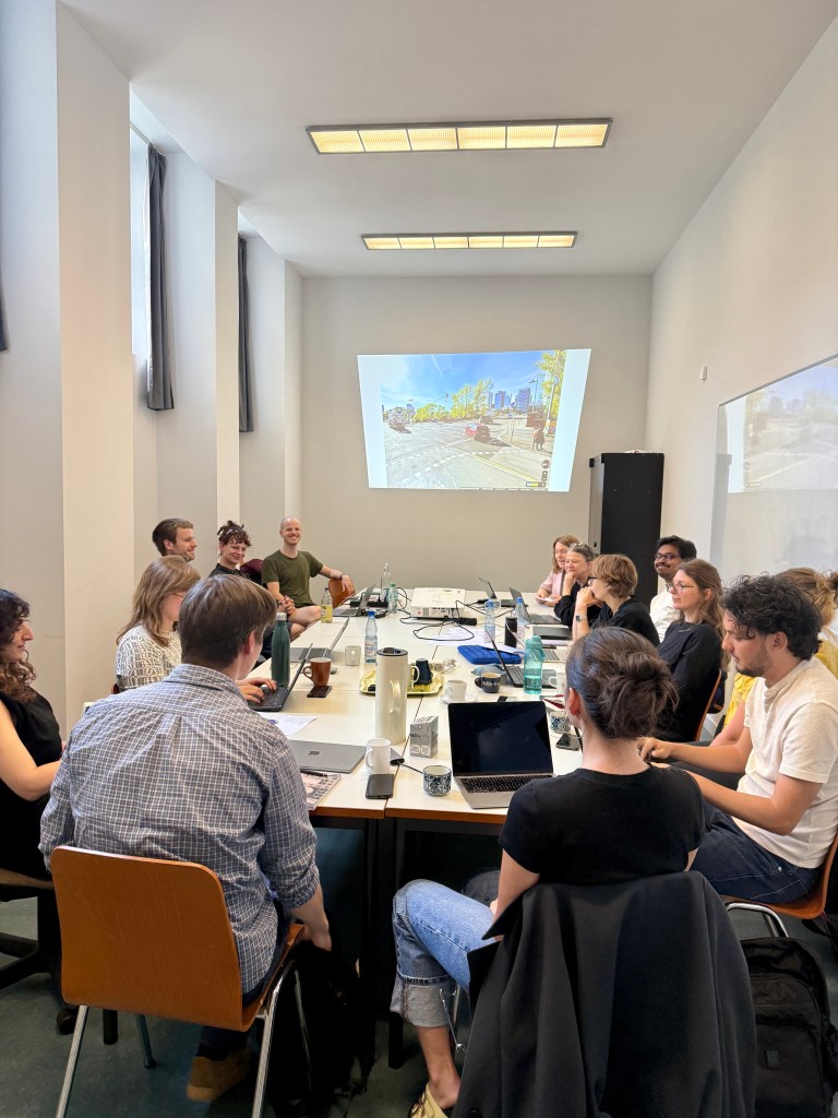 About a dozen people sit at a table in a narrow but brightly lit room, looking at laptops and an image of a city intersection projected onto the wall.