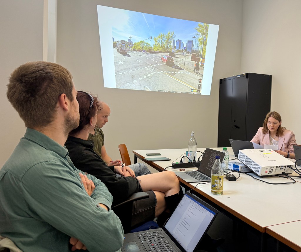 Several people sit at a table, looking at laptops and an image of a city intersection projected onto the wall.