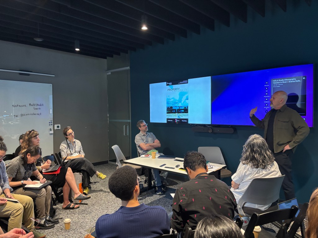 People sitting and standing in a conference room, listening to one person speak. The poster of the event is displayed on a large TV screen.