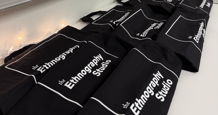 A table with neat stacks of black tote bags with the Ethnography Studio logo.