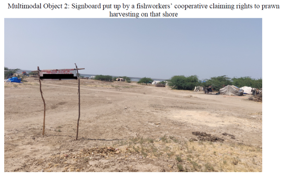 A dry dirt landscape with trees and tents a ways back in front of a shore, with a grey-blue sky. In the foreground, a sign is suspended between two sticks. The caption reads, "Multimodal Object 2: Signboard put up by a fishworkers' cooperative claiming rights to prawn harvesting on that shore."