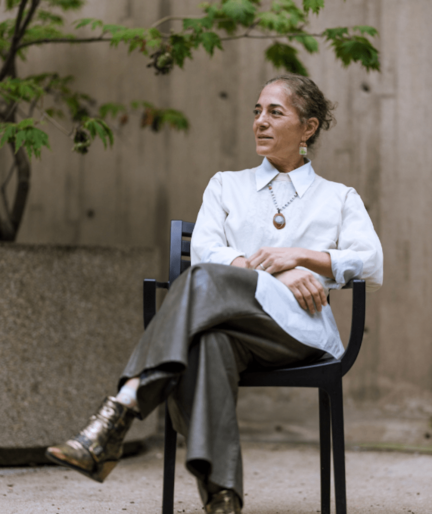 A person wearing a white shirt and black pants sits in a chair, legs crossed, and hair pulled back. There is a tree branch in the background.