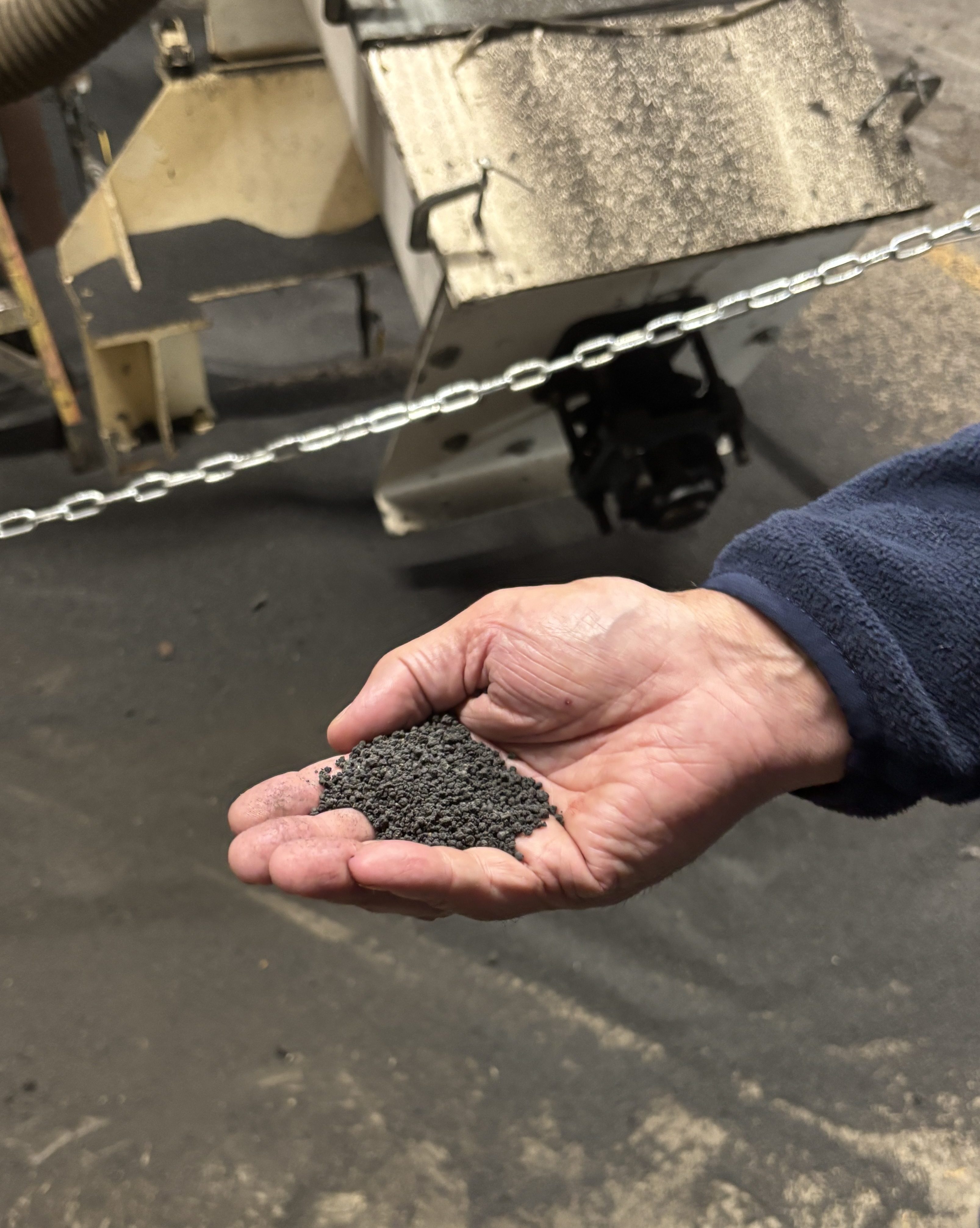 A hand holds small brown pellets in front of a metal machine with a chute.